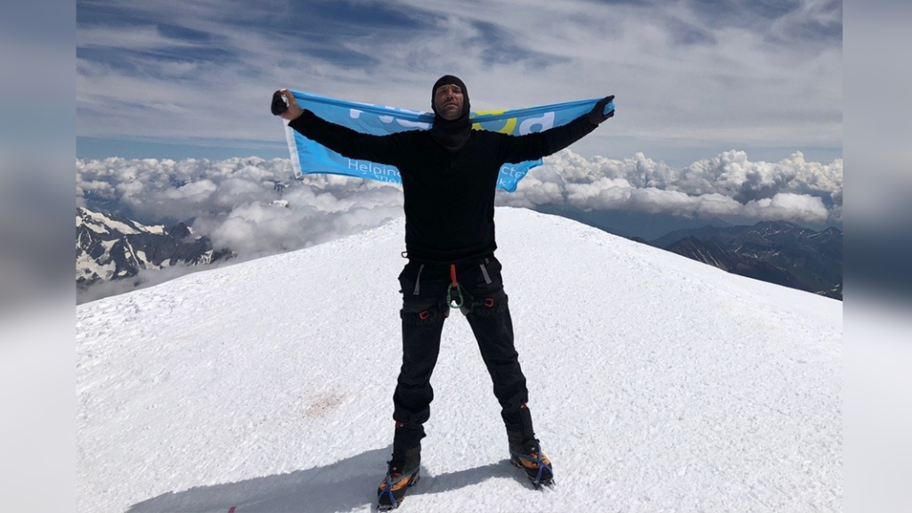 Calum Best standing on snow on top of Mont Blanc mountain with  blue Nacoa charity banner