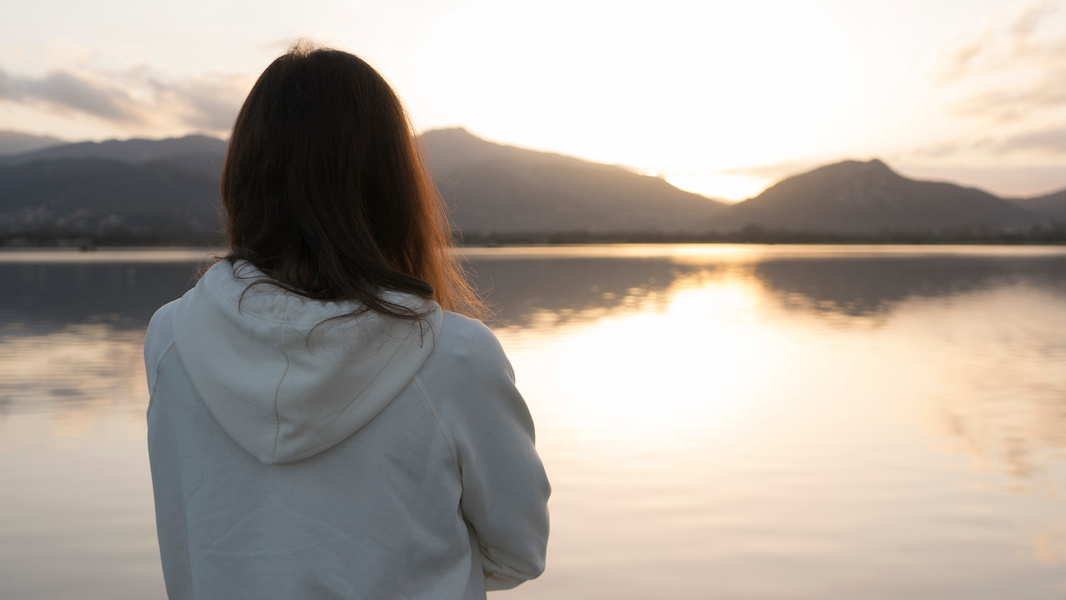 Young woman seen from behind looks at the lake at sunset reflecting on the impact of her mother's drinking and depression. Sarah says 'don't blame yourself for your parent's behaviour."